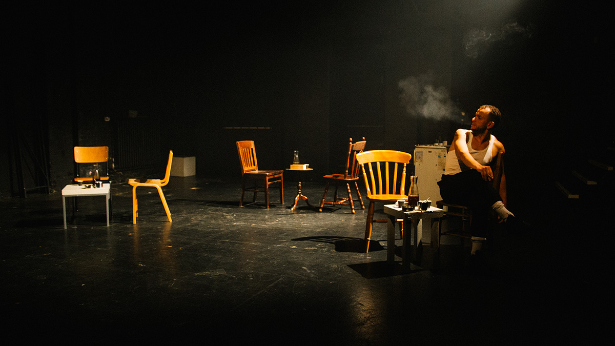 A man sits on a chair on the right of a dimly lit stage. Across the rest of the stage sits six other wooden chairs and tables.